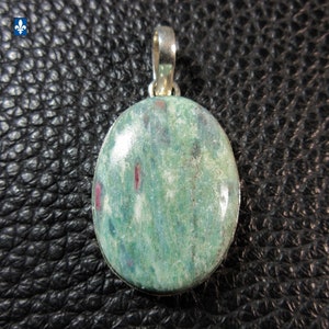 May include: An oval-shaped pendant featuring a polished green stone with visible white and dark mineral inclusions and distinct magenta-coloured flecks. The stone is set in a smooth silver-toned metal frame with a matching silver-toned bail at the top.