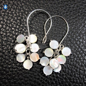 Peut inclure: Une paire de boucles d'oreilles pendantes argentées. Chaque boucle d'oreille présente un groupe de disques ronds nacrés irisés. Les boucles d'oreilles ont une fermeture à crochet de berger. Sur une surface noire texturée.