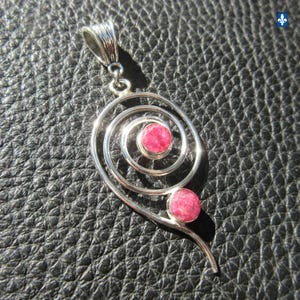 Könnte beinhalten: Ein silberner Anhänger mit einem spiralförmigen Drahtdesign und zwei runden, facettierten rosa Steinen, die in den Metallrahmen eingefasst sind. Der Anhänger verfügt über eine strukturierte Öse zur Befestigung und eine polierte Metalloberfläche.