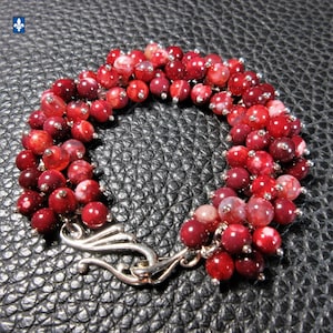 May include: A beaded bracelet featuring deep red and white speckled beads. The bracelet is secured with a silver-toned clasp. The beads are strung together, creating a curved shape. The bracelet is displayed on a textured black surface.