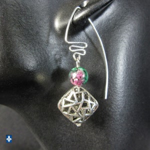 May include: Silver dangle earrings with a geometric design. Each earring has an openwork diamond-shaped charm, a round bead with pink and green detailing, and a decorative wire element. The earrings are displayed against a dark backdrop.