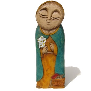 Op de afbeelding: Handgesneden houten beeldje van een persoon met gesloten ogen, gekleed in een turquoise gewaad en een boeket witte bloemen vasthoudend. Het beeldje staat op een houten voet met een mand.