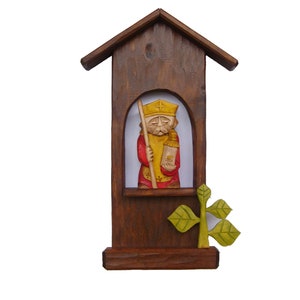 May include: A wooden shrine featuring a carved figure of a saint holding a staff and a book. The shrine is painted in shades of brown, red, and yellow. A small green plant is attached to the base of the shrine.