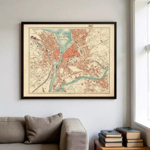 Peut inclure: Carte ancienne encadrée de Genève, Suisse, dans un cadre noir. La carte présente un plan détaillé de la ville avec des tons rouges et beiges, et des éléments bleus représentant l'eau. Le texte "Plan Genève" est visible en haut. La carte est affichée sur un mur blanc.