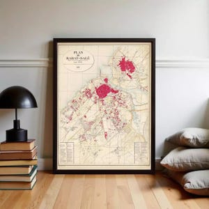 Può includere: Mappa vintage incorniciata di Rabat-Salé, Marocco, con una dettagliata rappresentazione cartografica in toni rossi e beige. La mappa, intitolata "Plan de Rabat-Salé", è in una cornice nera, appoggiata a un muro. A sinistra una lampada nera e libri.