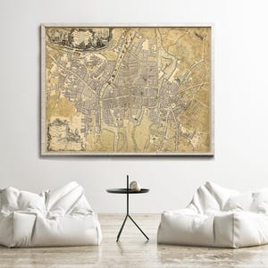 May include: A framed antique map print of a city, with detailed streets and buildings in shades of brown, beige, and grey. The print is displayed above two white bean bag chairs. The frame is light wood.