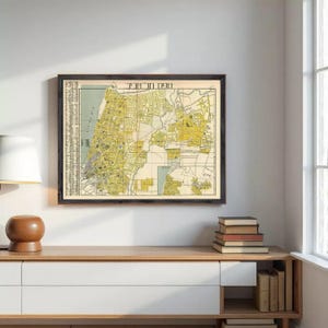 Puede incluir: Mapa vintage enmarcado de una ciudad, con un plano detallado de las calles en tonos amarillos y beige. El mapa está encerrado en un marco de madera oscura, exhibido en un mueble de madera clara. También se ven una lámpara y libros.