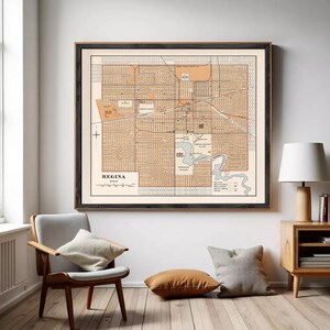 May include: A framed vintage map of Regina, Saskatchewan, Canada, in shades of tan and cream. The map features a detailed street grid, a river, and the city's name at the bottom. The frame is black, and the map is displayed on a white wall.