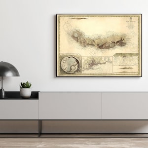 May include: A framed vintage map print in a black frame. The map has detailed illustrations and text, with a sepia colour palette. The print is displayed above a modern grey cabinet with a black metal frame, a black lamp, and a potted plant.