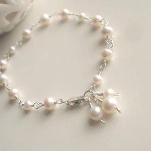 May include: A delicate bracelet featuring small, round, white freshwater pearls linked together with thin silver-toned wire. The bracelet includes a silver-toned clasp with three additional pearl charms dangling from the end near the closure.