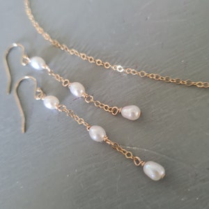 May include: A gold-coloured jewellery set comprising drop earrings and a necklace. The earrings and necklace feature white, teardrop-shaped pearls. The earrings have a hook fastening. The necklace is a delicate chain.