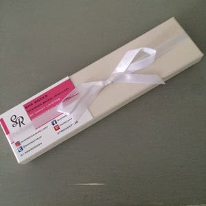 May include: A white box with a white ribbon tied in a bow. The box has a pink label with the text "SHE ROCKS DESIGNER BRIDAL JEWELLERY BY NANCY LAVERGE" and contact information for the company.