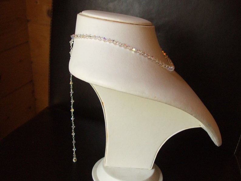 May include: A white crystal necklace with a long, dangling strand of crystals. The necklace is displayed on a white mannequin bust.