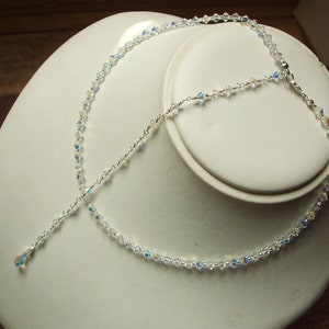 May include: A delicate silver necklace with a crystal pendant and a crystal chain. The necklace is displayed on a white mannequin bust.