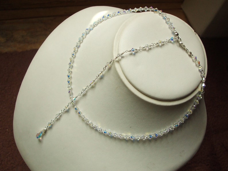 May include: A delicate silver necklace with a crystal pendant and a crystal chain. The necklace is displayed on a white mannequin bust.