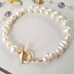 May include: A bracelet composed of white, irregularly shaped freshwater pearls strung together, featuring a gold-toned metal toggle clasp with a circular ring and a T-bar closure mechanism at the front.
