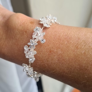 May include: A silver bracelet with a floral design. The bracelet features clear, sparkling crystals and small flower-shaped accents. The bracelet is worn on a wrist and appears to be made of metal.