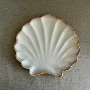May include: A scallop shell-shaped ceramic plate featuring a glossy white finish with subtle pinkish-purple gradients along the scalloped edges and a thin, defined brown rim outlining the entire perimeter of the dish.
