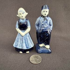 May include: Two ceramic figurines of a Dutch couple, hand-painted in shades of blue and white. The woman wears a traditional dress and bonnet, while the man is in a uniform. A quarter coin is next to the figurines.
