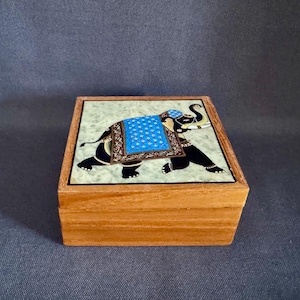 May include: Wooden box with a decorative elephant design on the lid. The elephant is black with a blue blanket and intricate detailing. The box has a light green background and a natural wood finish.