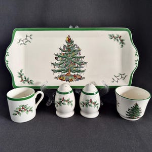 May include: A Christmas-themed serving set featuring a rectangular platter, a mug, a bowl, and two salt and pepper shakers. The set has a white background with a green border and holly accents. The platter features a Christmas tree design.