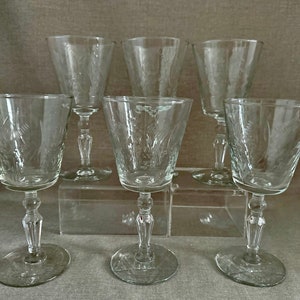 May include: Set of six clear, etched glass goblets. Each goblet features a flared bowl, a detailed stem, and a round base. The etched design appears to be a floral or leaf pattern. The glasses are arranged on a clear surface.