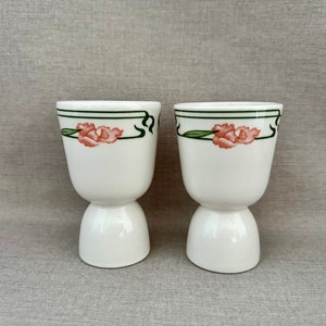 May include: Two white ceramic egg cups featuring a delicate hand-painted design of pink flowers with green leaves and a thin dark green border line encircling the upper rim of each piece, showcasing a smooth finish and a pedestal base.