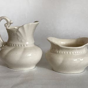May include: A cream-colored ceramic creamer pitcher and a matching open sugar bowl. Both pieces feature a rounded, bulbous body with a subtle vertical ribbed texture, a decorative beaded band around the midsection, and a scalloped rim edge.