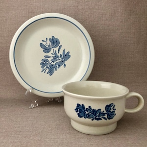 May include: Cream-colored teacup and saucer set with blue floral design. The saucer has a blue border and a floral motif. The teacup features a matching floral pattern. A classic, vintage-style tea set.