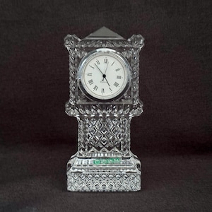 May include: A crystal clock with a clear face and black Roman numeral hour markers. The clock is in the shape of a tower and has a diamond pattern.
