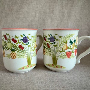 May include: Two white ceramic mugs featuring a colorful illustration of a fruit tree with green leaves, red cherries, purple grapes, yellow pineapples, pears, and strawberries. Each mug has a curved handle and a thin orange rim at the top.