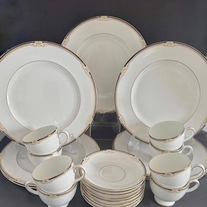 May include: A formal white dinnerware set with gold trim and decorative accents. The set includes large plates, saucers, and teacups. The teacups are stacked, and the saucers are neatly arranged. The plates are arranged in the background.