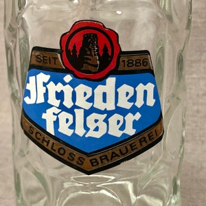 May include: Clear glass beer stein with a blue and white label. The label features the words "Friedenfelser Schloss Brauerei" and the year "1886". A red seal with a castle image is at the top of the label.