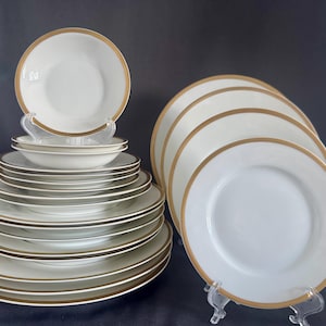 May include: A collection of white ceramic dinnerware featuring a simple gold rim. The set includes a stack of various sized plates and bowls, along with two individual plates displayed upright on clear plastic stands to show the gold trim detail.
