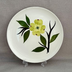 May include: A round white ceramic dinner plate featuring a hand-painted design of a single yellow flower with dark green leaves and black stems. The flower petals have small red accents at the tips, and the plate has a smooth, glossy finish.
