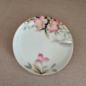 May include: A white porcelain saucer with a floral design featuring pink flowers and green leaves. The saucer has a small handle on the side.