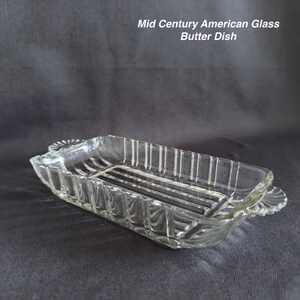 May include: A clear glass butter dish with a rectangular shape, featuring vertical ribbed sides and decorative fan-shaped handles on each end. The text Mid Century American Glass Butter Dish is printed in white in the upper right corner of the frame.