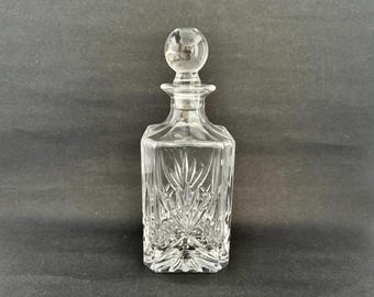 May include: A clear glass decanter with a crystal-cut design and a round stopper. The decanter has a square base and a tall, slender neck.