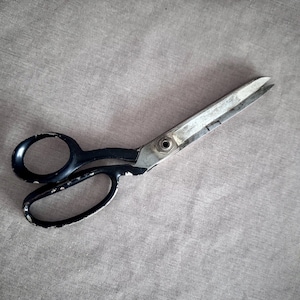 May include: A pair of vintage scissors with black handles and silver blades, resting on a gray fabric surface. The scissors show signs of wear, suggesting they are well-used. Ideal for sewing and crafting projects.