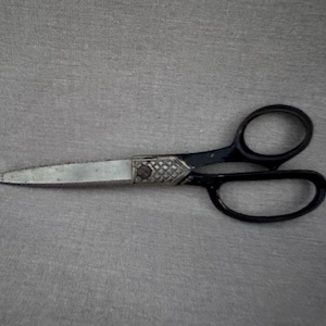 May include: A pair of vintage scissors with black handles and silver blades. The scissors have a decorative silver and patterned detail at the pivot point. The scissors are lying on a gray surface.