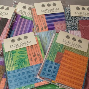 May include: A collection of Paste Papers Scrap Packs, each with a variety of colorful, patterned papers. The packs feature papers with stripes, woven textures, and floral designs in shades of orange, blue, green, and purple. Each pack has a label with the text "PASTE PAPERS SCRAP PACK".