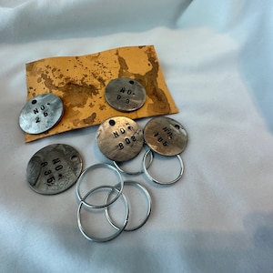 May include: Five silver metal tags with black numbers on them. The tags are attached to four silver key rings. The tags read "NO. 25", "NO. 98", "NO. 803", "NO. 106", and "NO. 636".