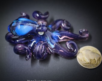 May include: A small glass octopus figurine with purple and blue iridescent glass. The octopus has eight tentacles and one eye. The figurine is sitting on a dark surface next to a coin.