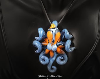 Puede incluir: Un collar con un colgante de vidrio en forma de pulpo con remolinos azules y naranjas. El pulpo tiene un cuerpo rayado en blanco y negro con un punto amarillo en la parte superior.