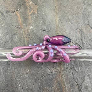May include: A purple glass octopus with blue accents is wrapped around a clear glass drinking straw. The octopus has a detailed design with curled tentacles and small dots.