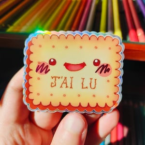 Peut inclure: Un autocollant holographique en forme de biscuit souriant. L'autocollant a un bord festonné et porte l'inscription "J'AI LU" en marron. Le biscuit a des joues et des yeux rouges.