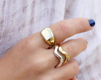 May include: A gold-toned ring with a smooth, rounded rectangular face and a second ring featuring a wavy, dual-tone design in gold and silver metal. Both rings have a polished, reflective finish and are worn on the fingers of a hand.