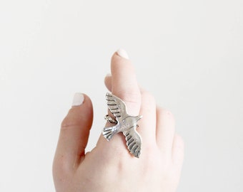 May include: A silver-colored ring in the shape of a bird in flight. The ring is worn on a finger, with the bird's wings outstretched. The ring is detailed with feathers and is set against a white background.