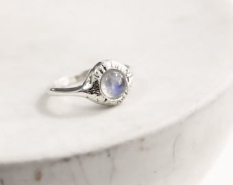 Könnte beinhalten: Ein Silberring mit einem runden, durchscheinenden Mondstein-Cabochon, der in eine texturierte Fassung mit Strahlenmuster eingefasst ist. Der Metallring hat eine polierte Oberfläche und der Stein zeigt ein dezentes blaues Schimmern.