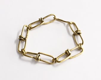 May include: A gold-toned metal chain bracelet composed of elongated rectangular links. Each link is connected to the next by a smaller, wrapped wire loop, creating a continuous circular design with a polished, metallic finish and a warm yellow hue.